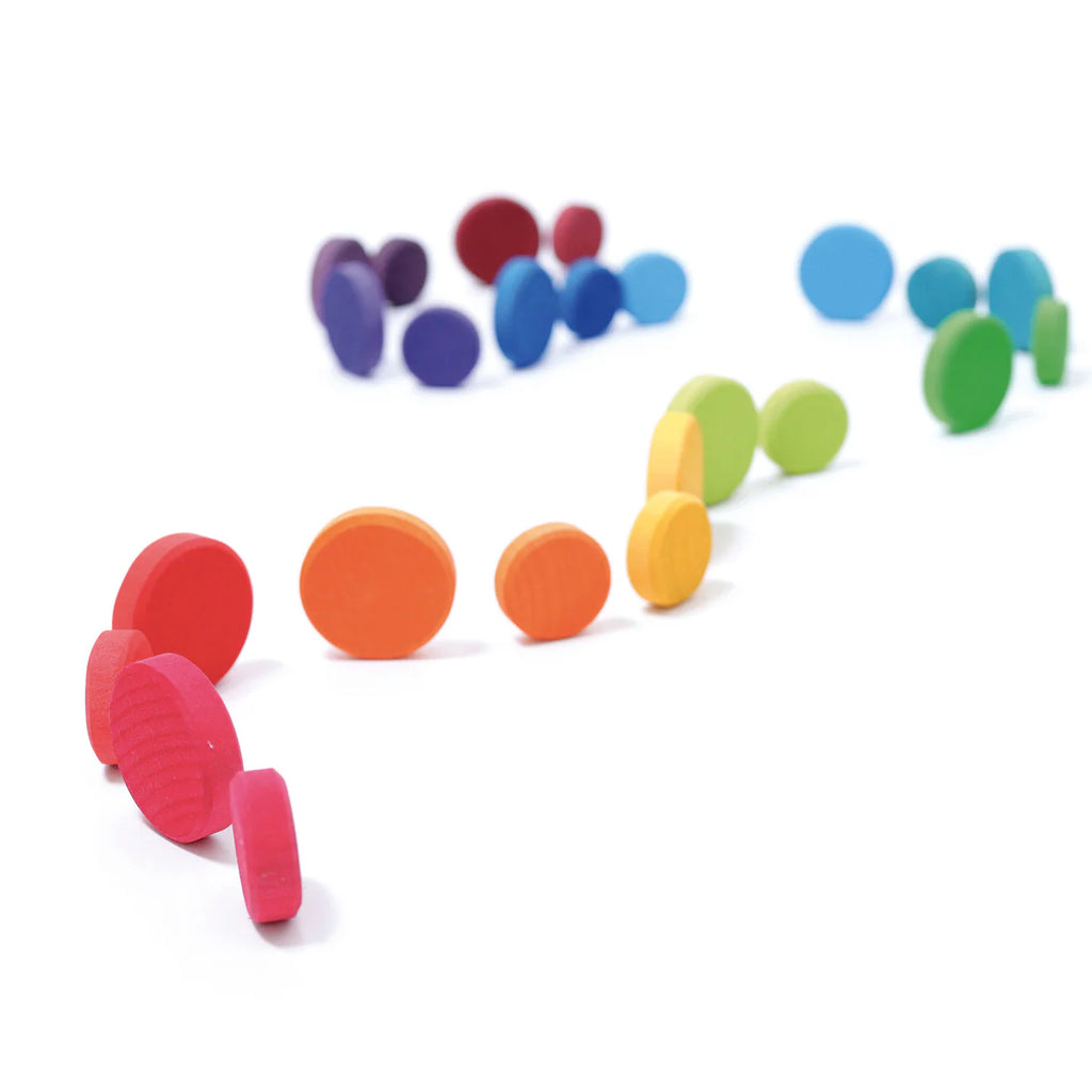 Grimm's Wooden Rainbow Coins (coming soon) feature vibrant wooden coins in red, orange, yellow, green, blue, and purple scattered on a white background—ideal tactile pieces for creative construction and imaginative play.