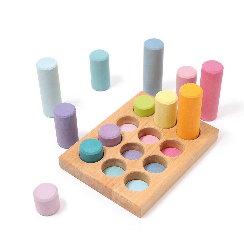 The Grimm's Stacking Game Small Pastel Rollers with various pastel-colored cylindrical blocks and corresponding circular slots enhances motor skills. Some construction pieces are placed in the slots, while others stand upright or lie around the wooden board, perfect for color sorting.