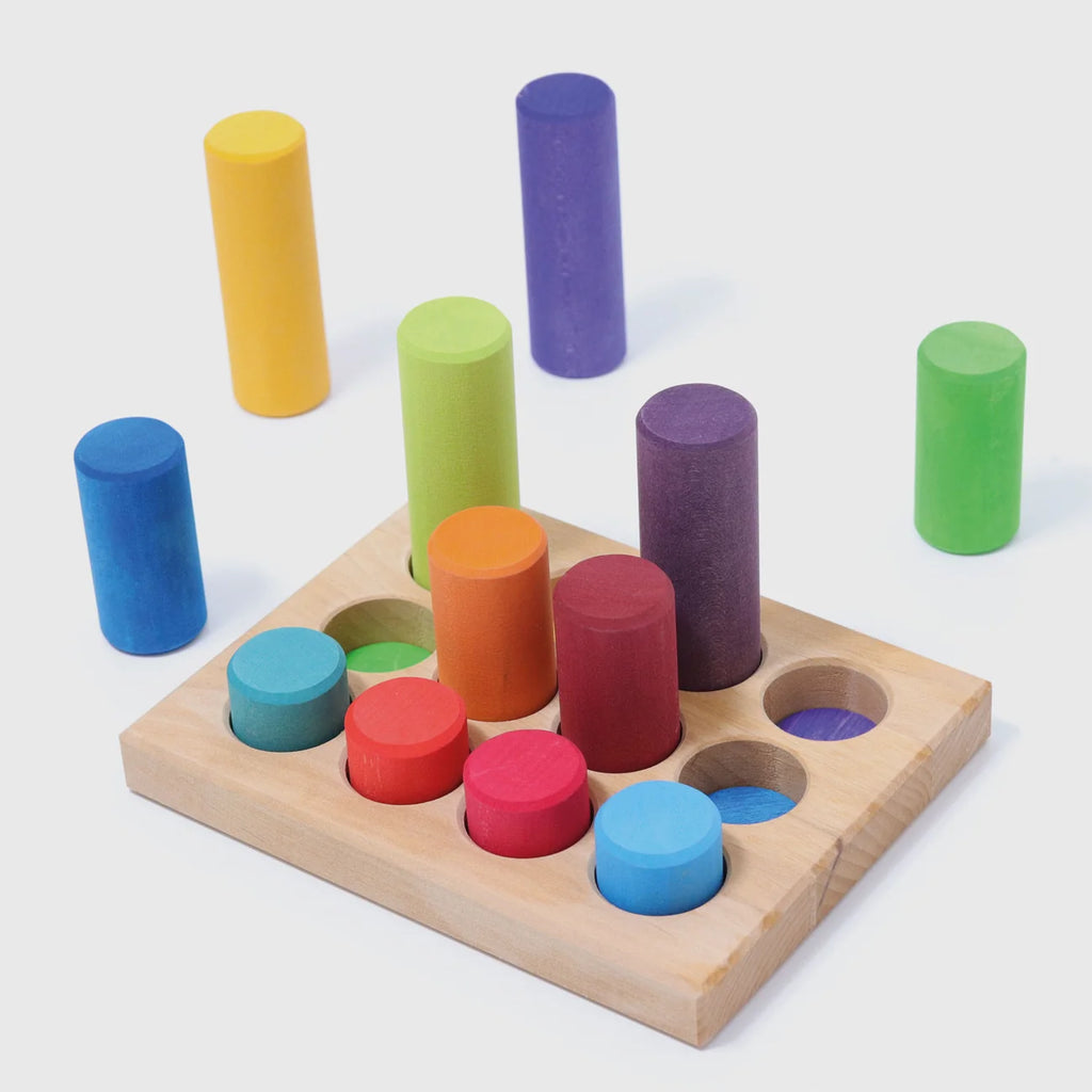 Grimm’s Stacking Game Small Rainbow Rollers featuring colorful wooden cylinders, with some placed in a perforated wooden base and others standing nearby.