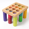 Grimm’s Stacking Game Small Rainbow Rollers featuring a wooden board with twelve circular holes, each containing a colorful cylinder that supports the board for color sorting and motor skills development.