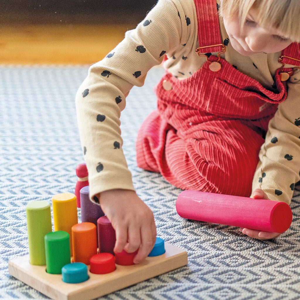 A child playing with a Grimm’s Stacking Game Small Rainbow Rollers wooden board and colorful, cylindrical stacking pieces in a color sorting game.