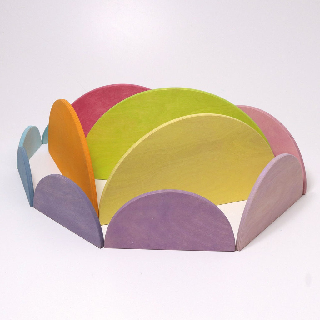 A colorful, wooden, Pastel Semi Circle Set arranged in a circle on a white background. Resembling modern architecture with its pastel shades of purple, blue, pink, yellow, green, and orange, this extra large creative puzzle forms a 3D rainbow pattern.