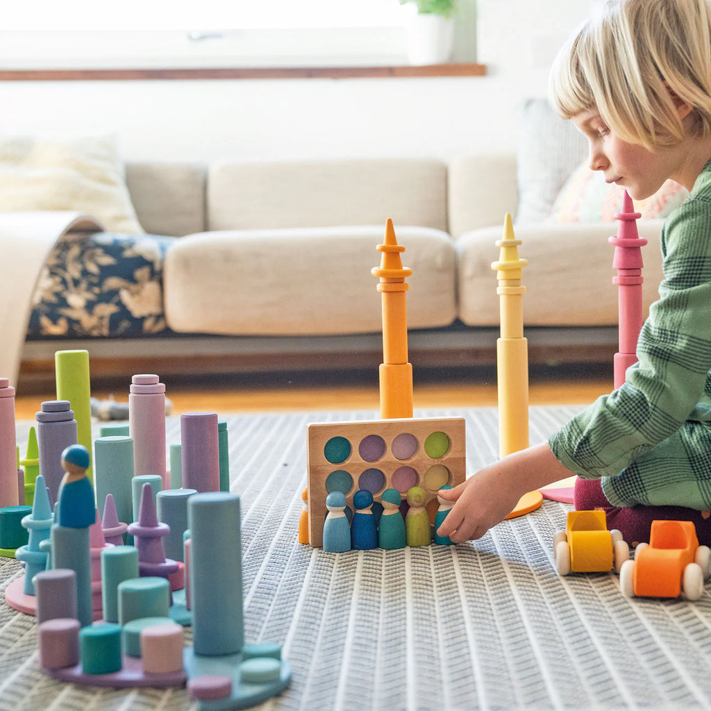 A young child in a green plaid shirt plays a stacking game on a carpet with Grimm's Large Building Rollers - Pastel (coming soon) and peg dolls, building motor skills. A beige couch with cushions is visible in the background.