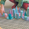 A child kneels on a patterned rug, developing motor skills while arranging Grimm's Large Building Rollers - Pastel (coming soon), colorful wooden cones and cylinders in soft pastel shades.
