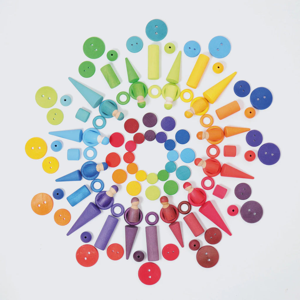 A vibrant mandala featuring Grimm's Wooden Rainbow Coins (coming soon), artfully arranged with tactile construction pieces—cones, rings, cylinders, stacking rollers, and peg dolls—in a rainbow gradient on a white background.