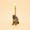 The Jaguar Cake Topper - Final Sale is a hand-painted ceramic figurine featuring a leopard wearing a blue party hat and carrying a lit yellow candle on its back, set against a plain beige background.
