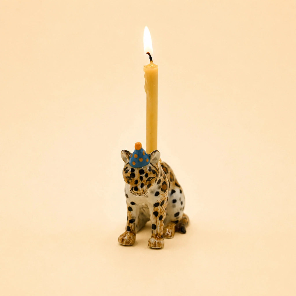 The Jaguar Cake Topper - Final Sale is a hand-painted ceramic figurine featuring a leopard wearing a blue party hat and carrying a lit yellow candle on its back, set against a plain beige background.
