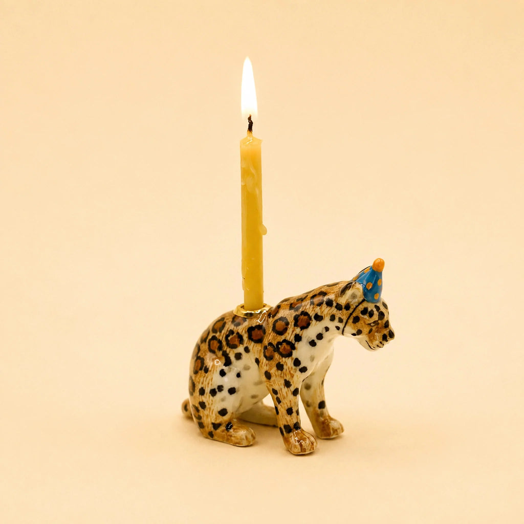 The Jaguar Cake Topper - Final Sale is a hand-painted ceramic figurine featuring a leopard sporting a small blue party hat and carrying a lit yellow candle on its back. Set against a simple beige background, this charming piece makes an ideal collectible keepsake or porcelain cake topper for any special celebration.