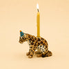 Celebrate in style with the Jaguar Cake Topper – Final Sale, a hand-painted ceramic figurine featuring a jaguar wearing a blue party hat and carrying a lit yellow candle on its back, showcased against a plain beige background.