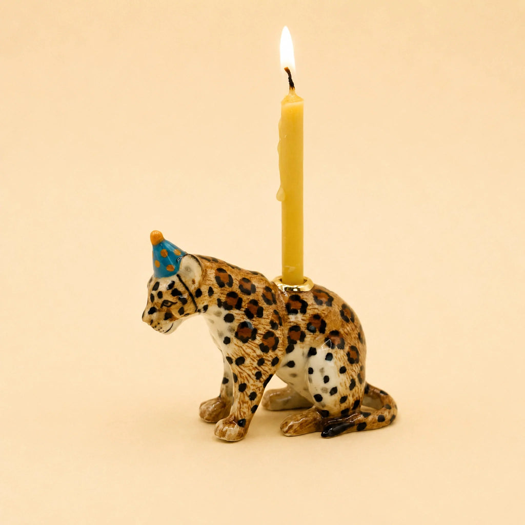 Celebrate in style with the Jaguar Cake Topper – Final Sale, a hand-painted ceramic figurine featuring a jaguar wearing a blue party hat and carrying a lit yellow candle on its back, showcased against a plain beige background.