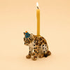 Introducing the Jaguar Cake Topper – Final Sale: this hand-painted figurine features a jaguar wearing a blue party hat with orange dots and carrying a lit yellow candle on its back, displayed against a plain beige background.