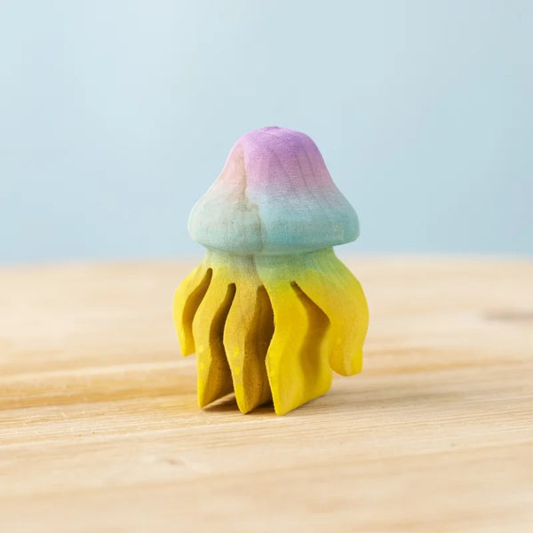 The Bumbu Handmade Wooden Jellyfish features a purple, blue, green, and yellow gradient and rests on a light wood surface with a pale blue background—an eco-friendly ocean toy ideal for Montessori play.