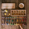 A child's feet visible next to an organized display of colorful Grapat Mix & Match cubes sorted by color in a wooden sorting box and spoons on a rustic wooden floor.