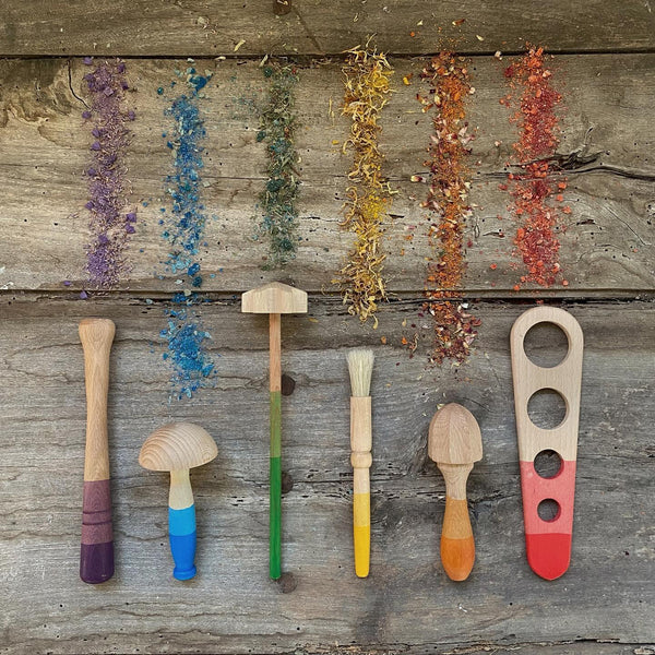 An assortment of colorful spices and herbs arranged linearly next to Grapat Tools, including a wooden mallet, paintbrush, and spoons, on a rustic wooden background.