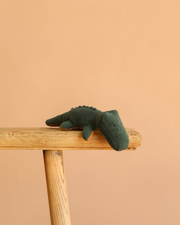 The Maileg Crocodile, Mini - Forest Green, a huggable plush toy, sits on the edge of a wooden bench with a plain peach background.