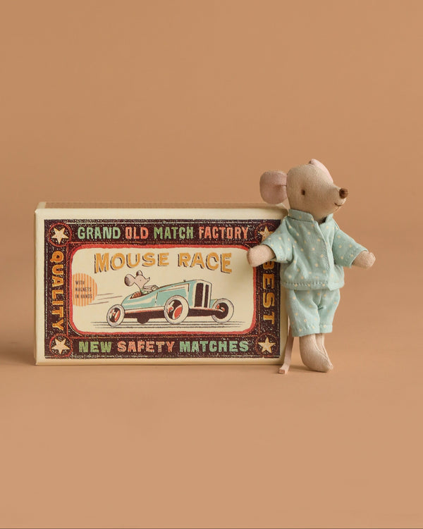 The Maileg Little Brother Mouse in blue pajamas leans against its vintage-style matchbox, which features a race car illustration and retro text, all set on soft fabric bedding with a charming beige background.