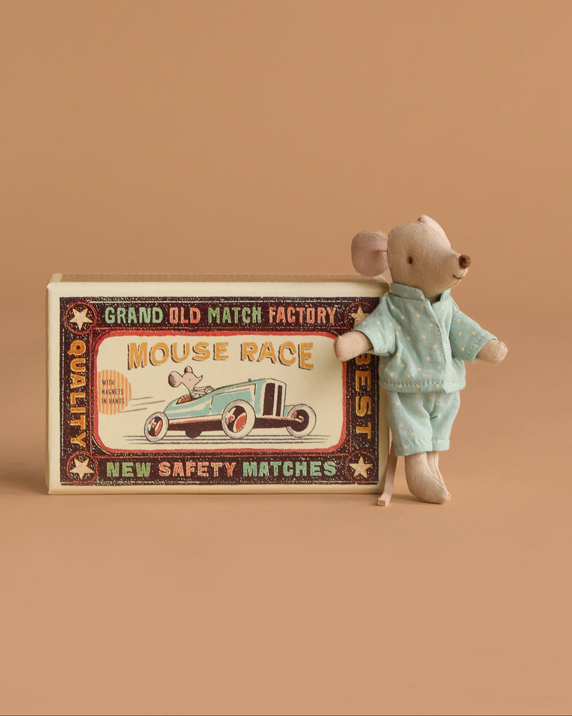 The Maileg Little Brother Mouse in blue pajamas leans against its vintage-style matchbox, which features a race car illustration and retro text, all set on soft fabric bedding with a charming beige background.