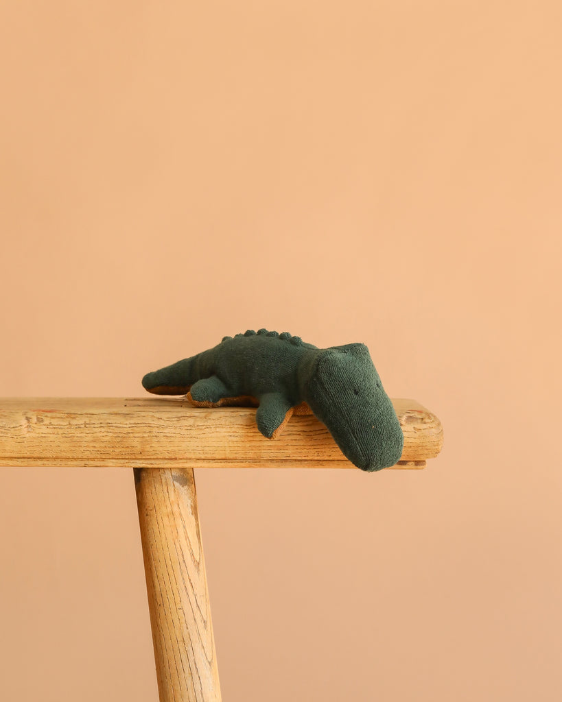 The Maileg Crocodile, Mini - Forest Green, a huggable plush toy, sits on the edge of a wooden bench with a plain peach background.