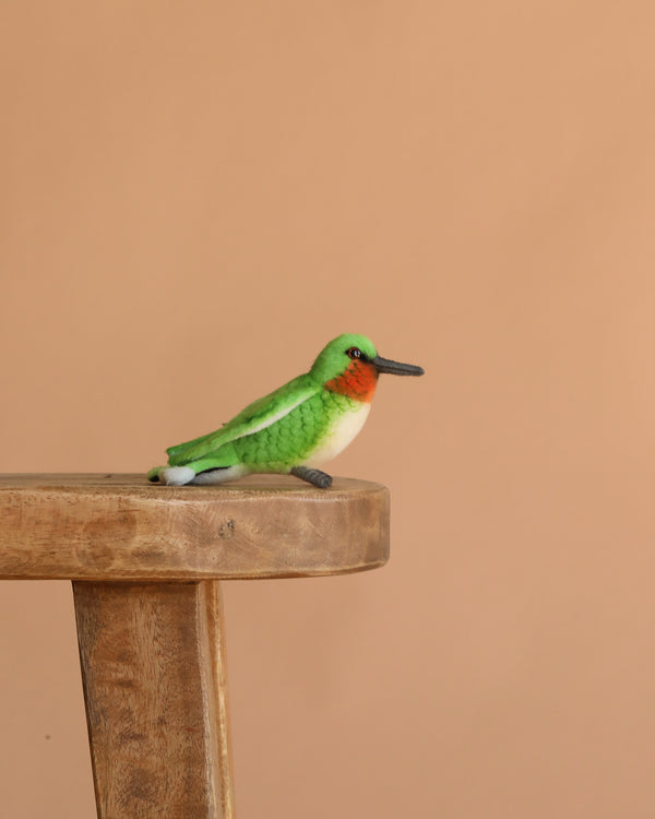 The Hummingbird Stuffed Animal, a small and colorful plush bird with green wings, a red throat, and a white belly from the HANSA animals collection, sits on a wooden stool against a plain beige background.