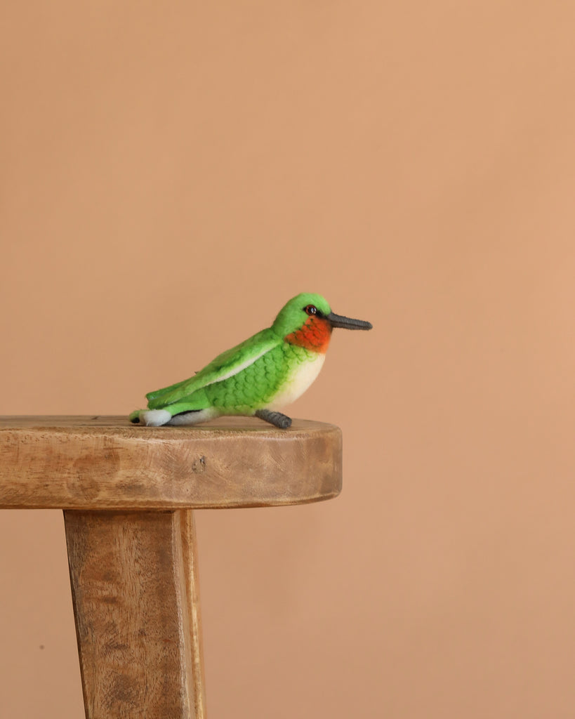 The Hummingbird Stuffed Animal, a small and colorful plush bird with green wings, a red throat, and a white belly from the HANSA animals collection, sits on a wooden stool against a plain beige background.