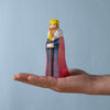 A hand holds the Bumbu Handmade Wooden King, a sustainable wooden toy with a yellow crown, red cape, and white collar with black spots, displayed against a plain light blue background.