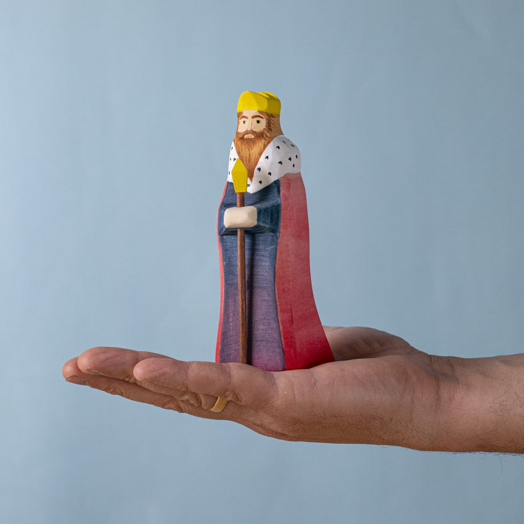 A hand holds the Bumbu Handmade Wooden King, a sustainable wooden toy with a yellow crown, red cape, and white collar with black spots, displayed against a plain light blue background.