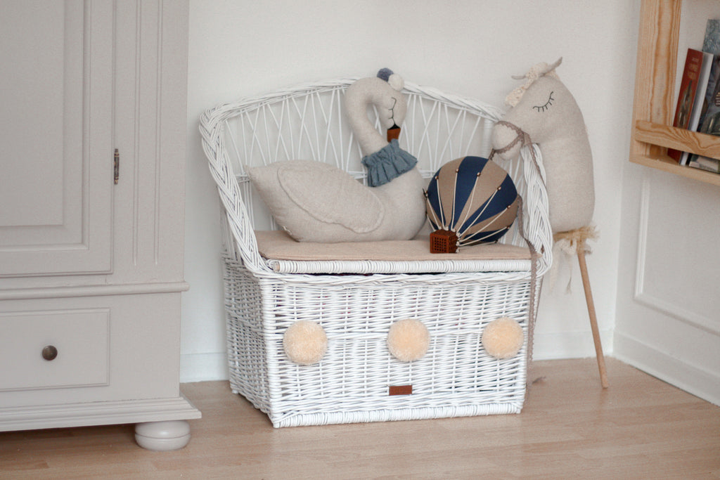 A white wicker bench with a cushion, decorated with a swan plush, llama doll, and the Hot Air Balloon - Light Brown / Navy Blue, sits in the corner of a children's room with light wood floors and neutral furniture.
