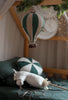 A cozy, softly lit bedroom features the Hot Air Balloon - Light Brown / Green decoration hanging above a bed with matching green bedding, a round pillow, and a plush unicorn. Greenery and string lights accent the wooden frame in this charming kids' room.