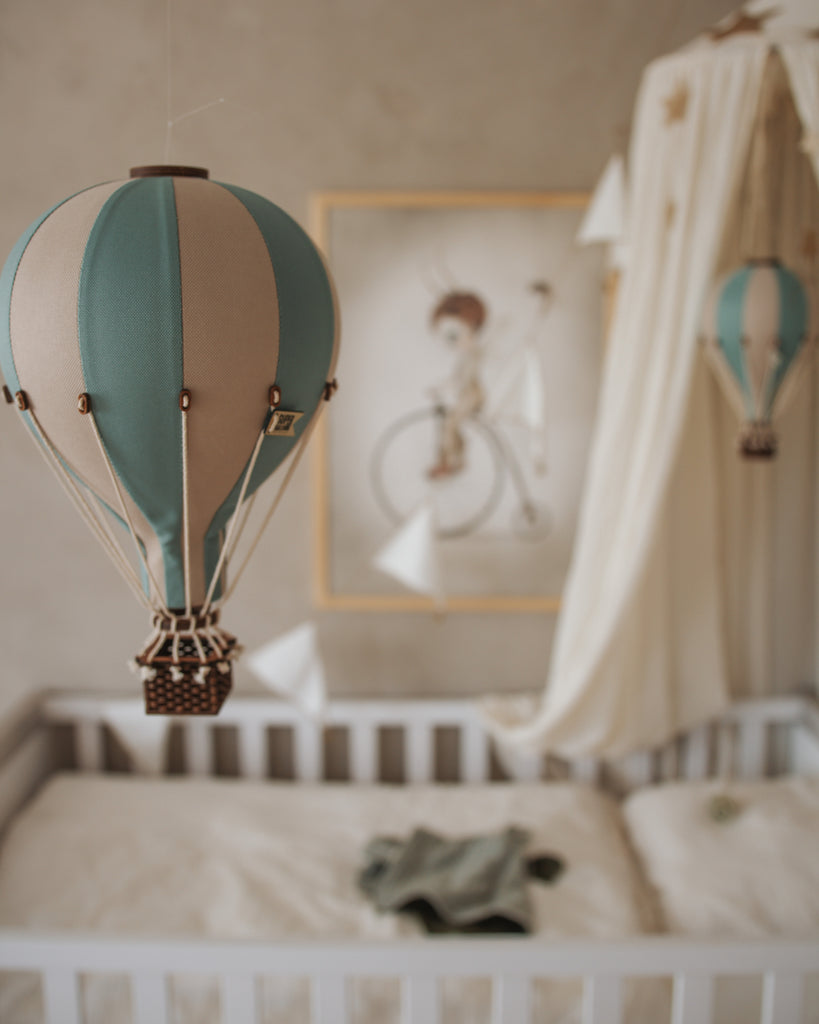 A nursery with a white crib, soft bedding, and a hanging Hot Air Balloon - Pastel Green decoration. In the background, a framed illustration of a child on a bicycle adds to the handmade children's room decor charm.