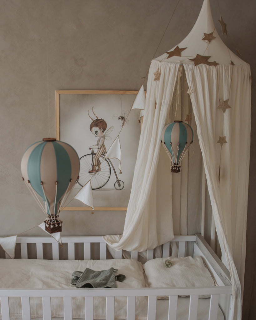 A cozy nursery corner featuring a white crib, cream canopy with gold stars, the Hot Air Balloon - Pastel Green ornament, and a framed child-on-bicycle illustration—ideal for anyone seeking handmade children's room decor.