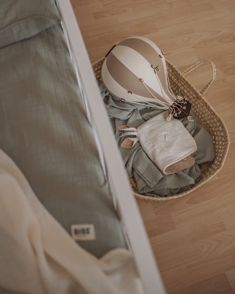 A wicker basket on a wooden floor holds folded baby clothes and the Hot Air Balloon - Gold, ideal for children's room decor, placed beside a bed with greenish-gray bedding.