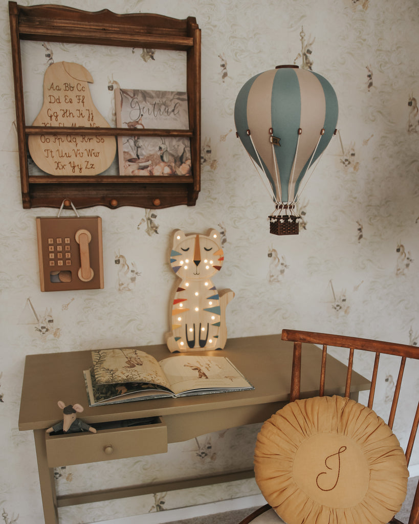A child's desk with an open storybook, cat-shaped lamp, and wooden chair. Above are a pastel green Hot Air Balloon decoration, an alphabet plaque, shelf signs, all set against floral wallpaper.