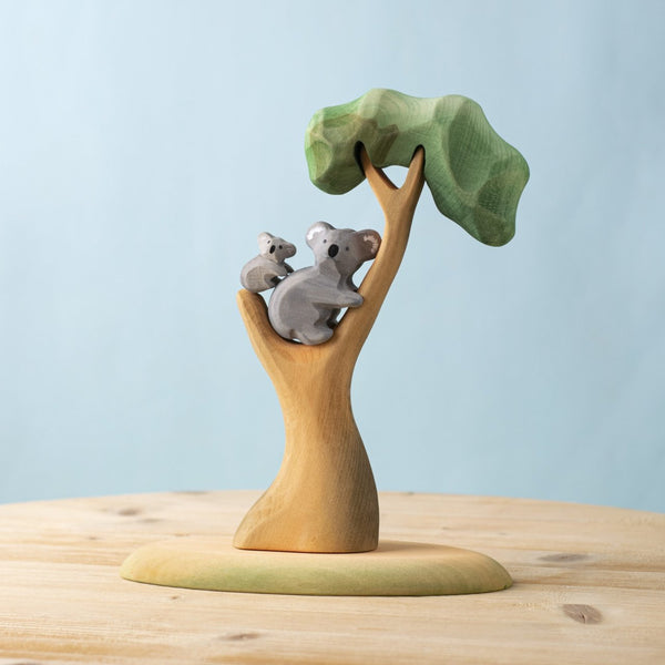 The Bumbu Handmade Wooden Koala Bears with Eucalyptus Tree Set features an adult and baby koala on green-leaved branches, displayed on a light wooden surface against a blue background.