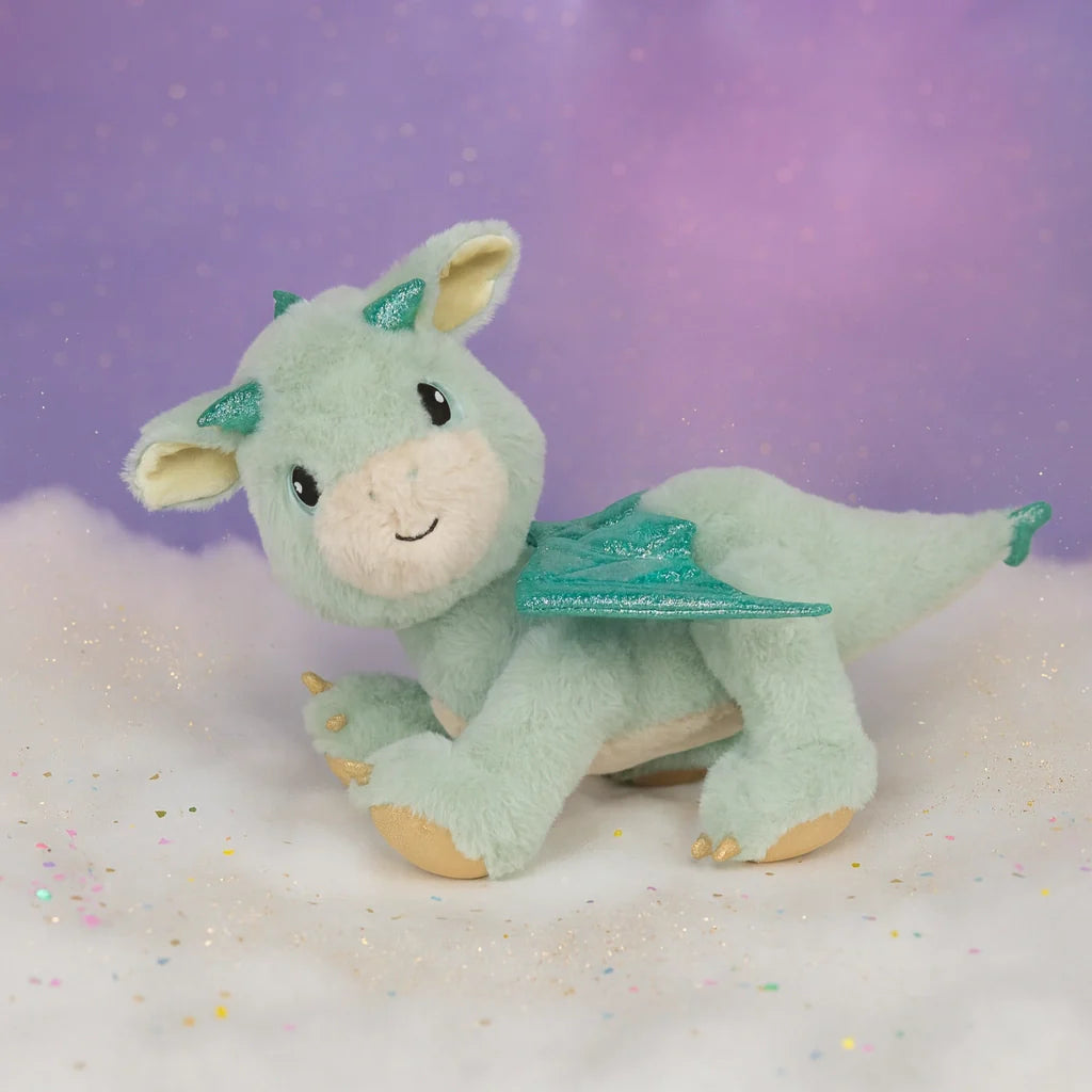The Olli Ella Dinkum Dragon - Blaze - Green is a plush dragon toy with sparkly teal wings and horns, a smiling face, cream accents on its belly and feet, and a magnetic mouth. It sits on a fluffy white surface against a starry purple background.