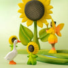 Wooden toys—including a goose, two sunflower people, the Bumbu Handmade Wooden Large Sunflower sculpture, and a fairy with orange wings—are arranged on a light green surface with a matching background, creating charming eco-friendly decor that highlights sustainable wood toy artistry.