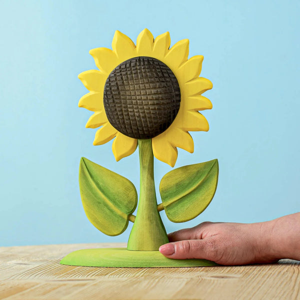 A hand holds the Bumbu Handmade Wooden Large Sunflower, featuring yellow petals, green leaves, and a textured dark brown center. It stands elegantly on a wooden surface against a light blue background—an exquisite piece of eco-friendly decor.