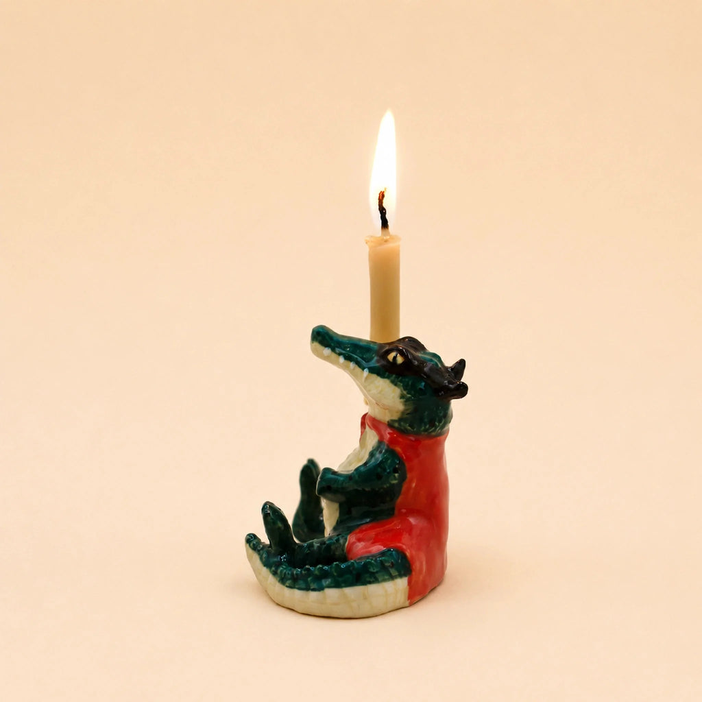The Later Gator Cake Topper - Final Sale features a charming ceramic crocodile figurine dressed in a red shirt and glasses, holding a lit candle on its head against a plain beige background, making it an ideal handcrafted keepsake and collectible birthday decoration.