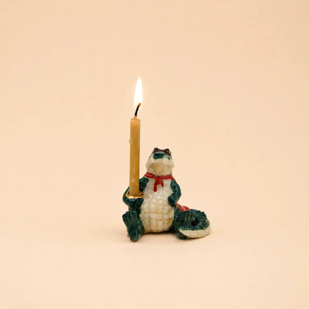 A handcrafted keepsake, the Later Gator Cake Topper features a ceramic alligator figurine wearing a red scarf and holding an upright lit candle in one hand, set against a plain beige background.