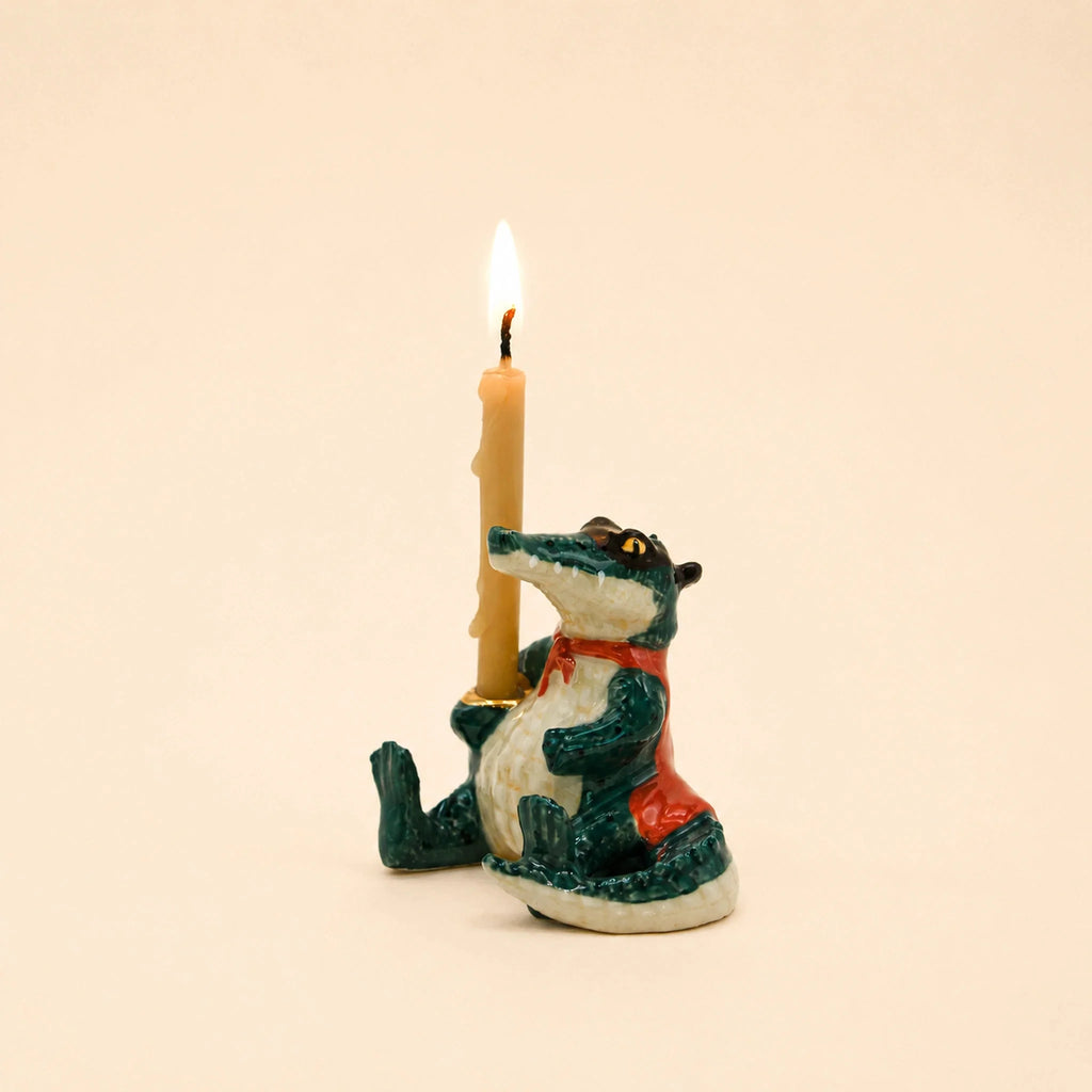 The Later Gator Cake Topper—a charming ceramic alligator figurine dressed in a red vest and holding an upright lit beige candle—rests against a simple backdrop, making it an adorable handcrafted keepsake and the perfect collectible birthday decoration.