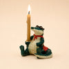 A small ceramic figurine named the Later Gator Cake Topper sits upright against a plain beige background, wearing a red cape and holding a lit yellow candle in one hand. Handcrafted and delightfully detailed, this keepsake makes a charming porcelain cake topper or collectible birthday decoration.