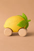 Lou The Lemon Baby Car, an eco-friendly, yellow lemon-shaped toy with green leaves and four wooden wheels, sits on a tan surface. "Oli & Carol" is embossed on the lemon.