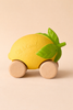 Lou The Lemon Baby Car is a yellow, lemon-shaped sensory toy with green leaves and four wooden wheels, shown on a beige background. "Oli & Carol" is embossed on the side of this eco-friendly baby car.