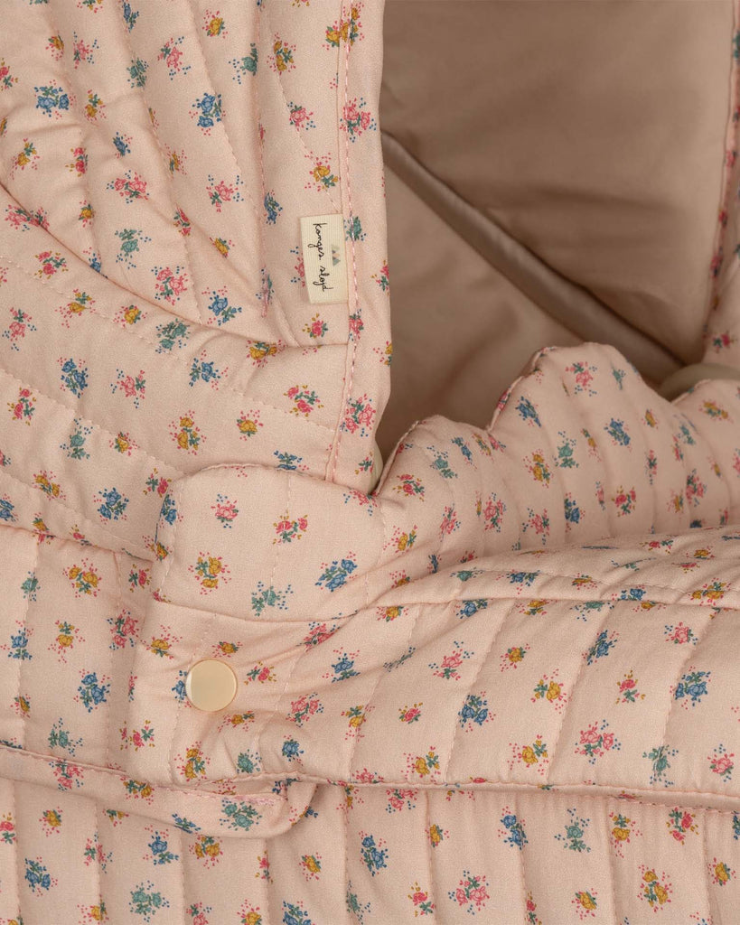 Close-up of the Doll Pram - Boomie Blush, featuring quilted fabric with a floral pattern in shades of blue, pink, and yellow on a beige background. Crafted from organic cotton, it is embellished with small buttons and a tag with indistinct text, showcasing a cozy and intricate design.