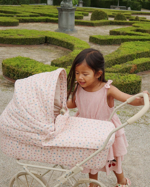 A young girl in a striped dress, smiling as she pushes the Doll Pram - Boomie Blush, crafted from recycled materials, through a garden with geometric hedges and pathways.