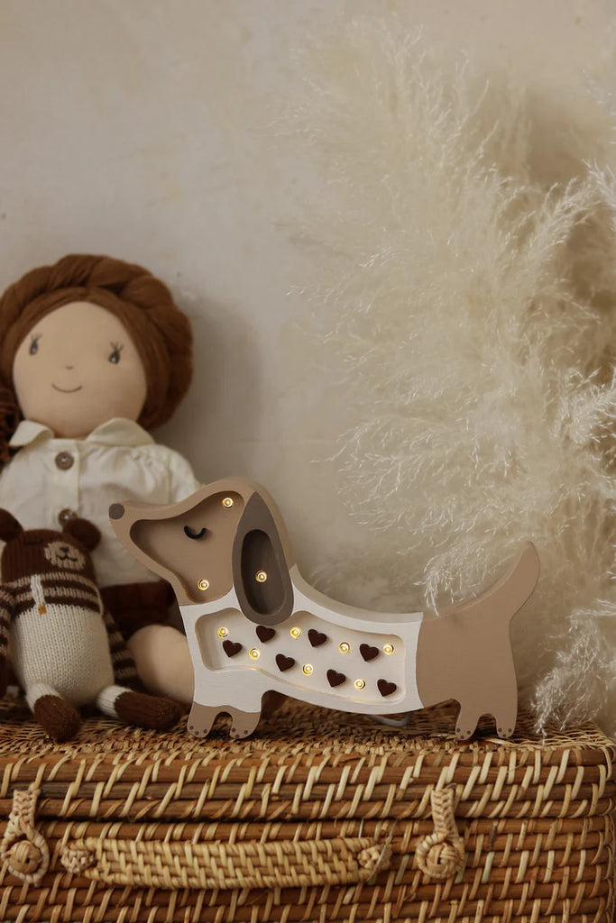 The Handmade Wooden Mini Lamp - Puppy, featuring heart-shaped cutouts, is a charming decorative piece perfect for nursery decor—ideal displayed on a basket beside cuddly toys with soft beige pampas grass in the background.