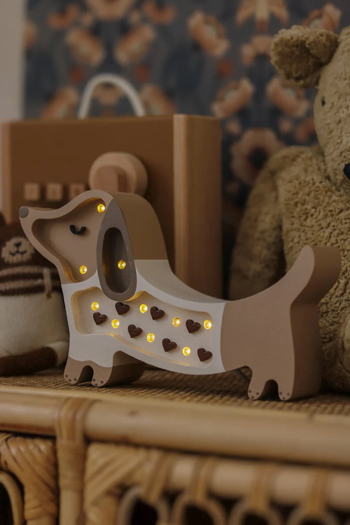 The Handmade Wooden Mini Lamp - Puppy, crafted from pine with glowing bulbs and heart cutouts, sits on a wicker shelf among plush toys—an ideal nursery decor piece for creating a cozy, softly lit room.