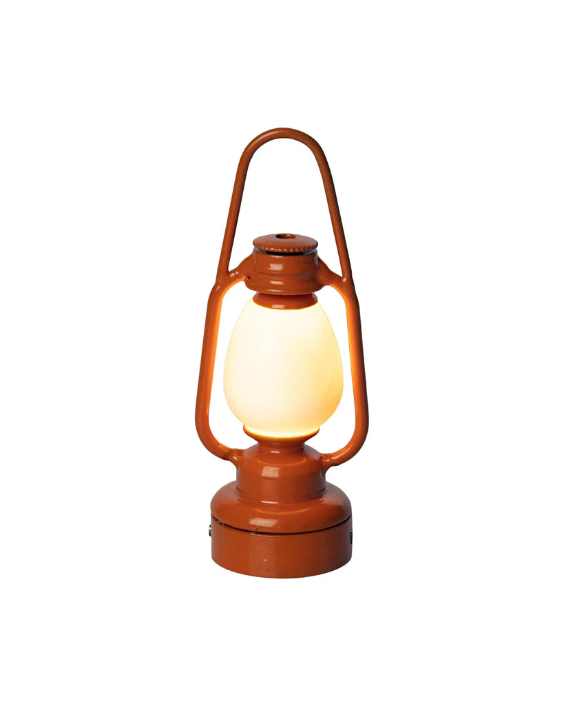 An orange Maileg Lantern with a glowing white light, featuring a rounded base and a metal handle loop. Resembling a miniature lantern, it has a classic, rustic design and is set against a plain white background.