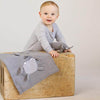 A cheerful baby with a light grey outfit leans on a large wooden box, holding an Organic Baby Lovey Blanket - Elephant and an organic cotton knit blanket, with a beige background.