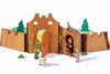 A wooden toy castle with attached trees is depicted, embodying the charm of handcrafted wooden toys. Various wooden figures, including a person with a green cloak and hat holding a bell, a dog, a woman and child, and a crowned figure, are artfully arranged around and inside the sustainably sourced jailhouse from Ostheimer Jailhouse.