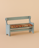 The Maileg Mouse Bench - Vintage Blue is a small, light blue wooden bench with a backrest and curved armrests. It has a vintage design and a striped orange, beige, and brown cushioned seat, displayed on a plain beige background.